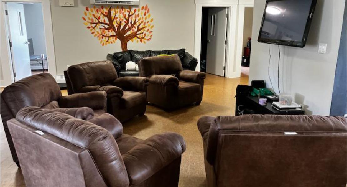 Living room with 5 brown recliners, TV, gaming systems, and light grey walls with a painting of an Oak tree. 