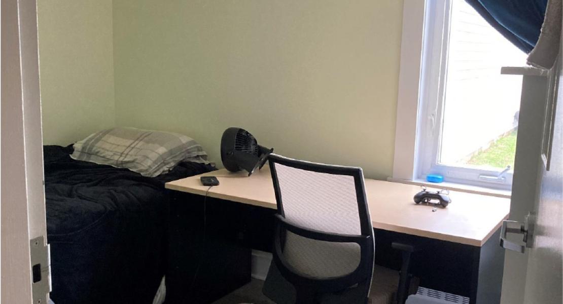 Cozy bedroom with light floors and light green walls, single bed, desk with fan and desk chair facing a small bright window. 