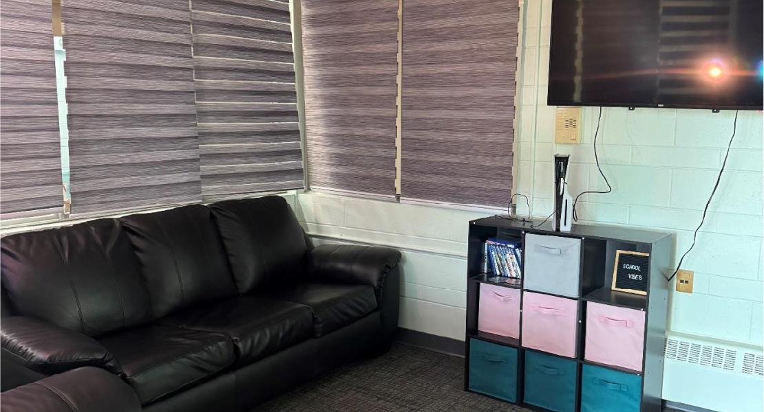 Light blue room with blinded windows, a black leather couch and chair, a shelf with colourful storage bins, a gaming system and a TV mounted on the wall 
