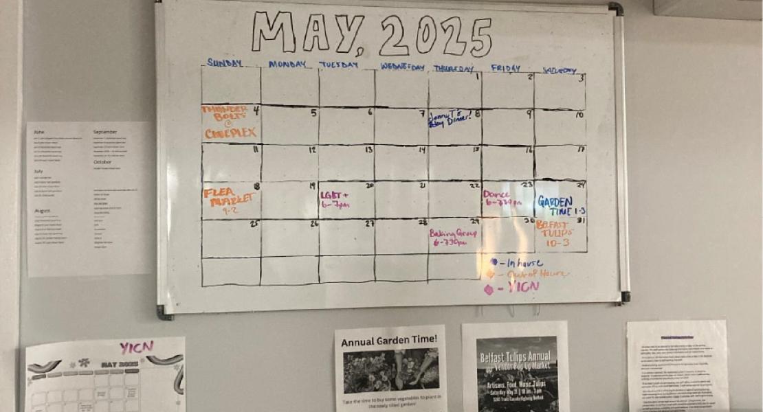 White board calendar with “May 2025” written at the top, hung on a light grey wall along with other event notices 