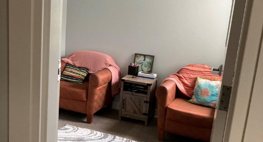 View of small room with two orange chairs and a small side table, patterned white and grey mat, grey walls and light brown floor, taken through a doorway 