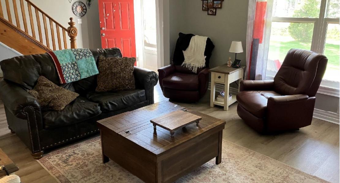 living room with large bright window, a couch and two reclining chairs next to an open doorway and staircase. 