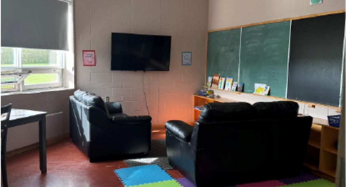 Small area with mounted TV, colourful foam mats, leather furniture, chalkboards on the walls and a large, bright window.