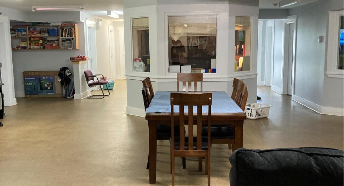 Large area with light flooring, black couches in foreground, small table in middle of room with four chairs, light blue walls with additional windowed room in the center. 