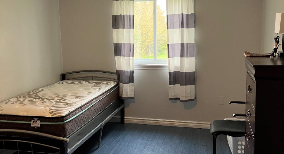 Bright room with dark floors, a single bed, chair and dresser, one window with striped black and white curtains 