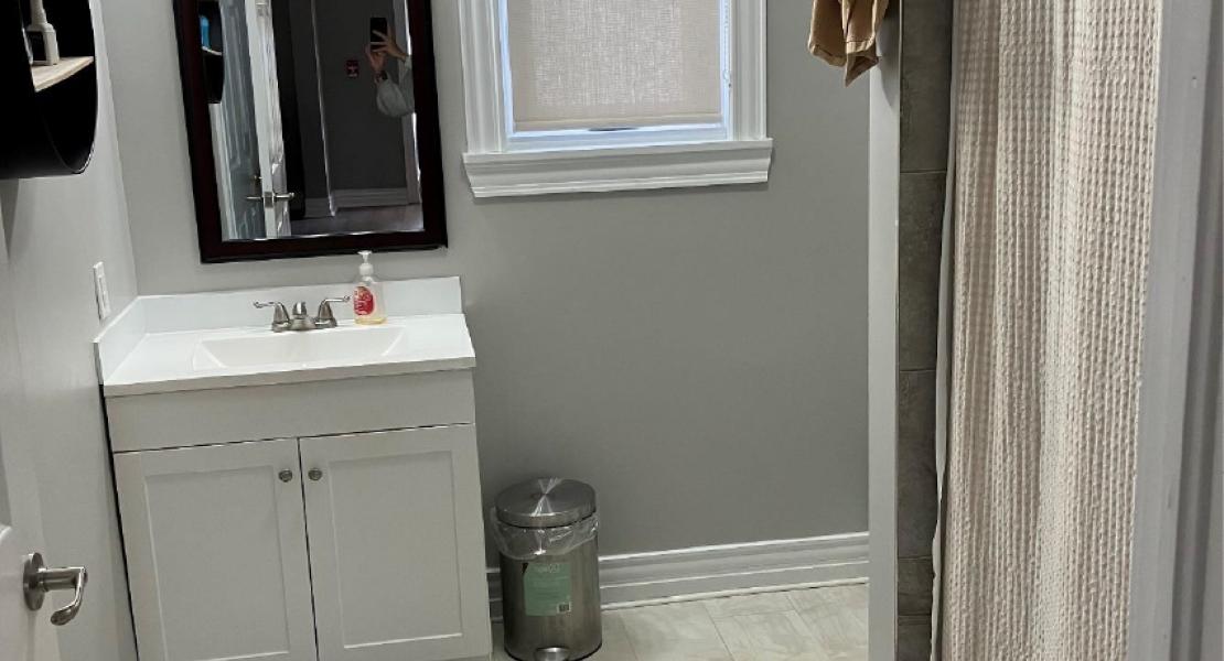  a bathroom with light flooring, a mirror on the wall over top of a vanity with sink and a stand-up shower with curtain. 