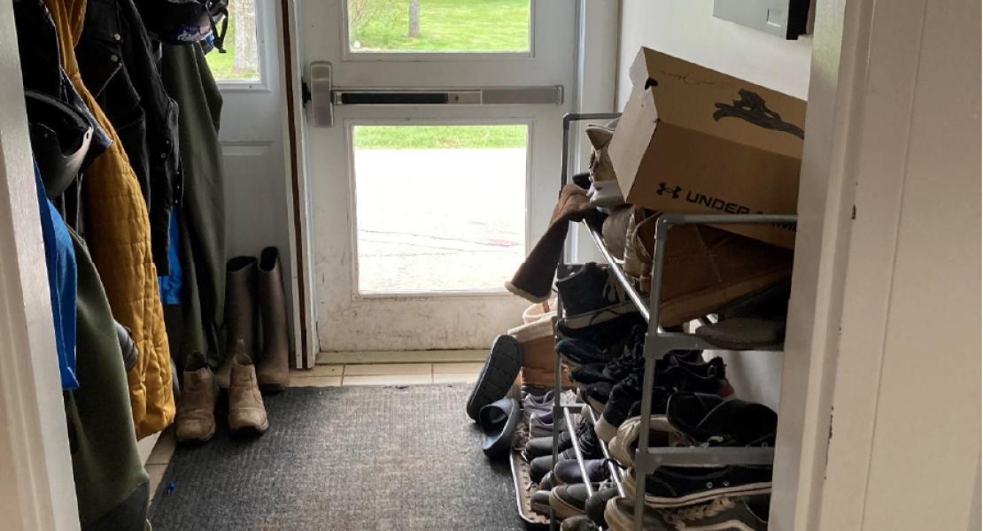 Tiled entryway with black mat, shoe rack filled with shoes with area for jackets. Large white door leading to outdoor area. 