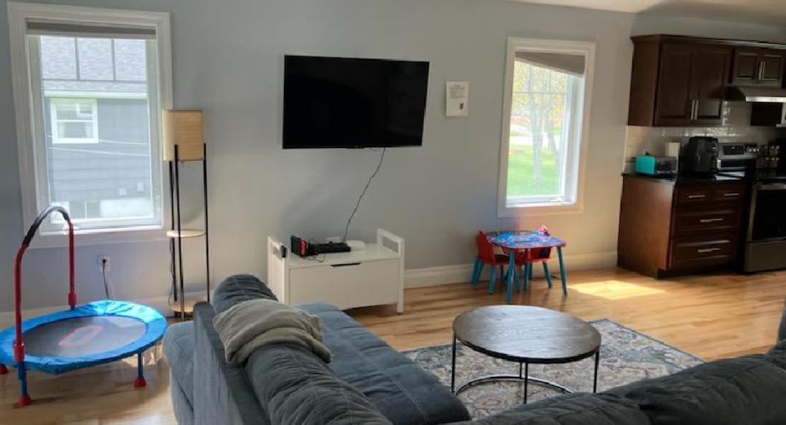 Living room with a dark couch, mounted TV, mini trampoline, kids’ table, and partial view of the kitchen. 