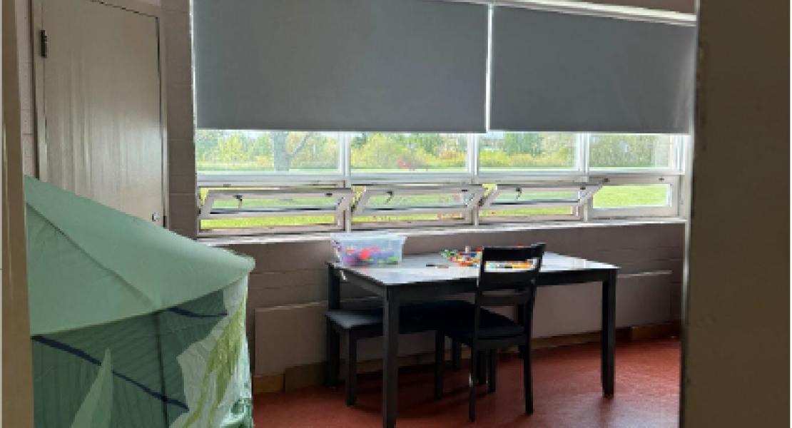 Small area with a large double window, desk with colorful blocks on top of it, green foam mats and a green play tent. 