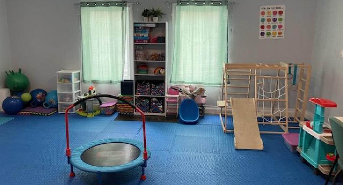 Children’s sensory room with blue flooring, mini trampoline, shelves of toys, climbing structure and two curtained windows
