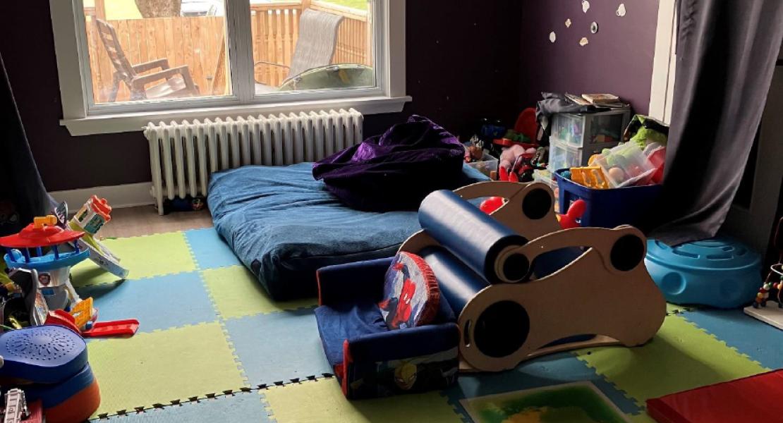 Sensory room with foam floor tiles, cushions, toys, and a large window showing a tree and outdoor seating area 