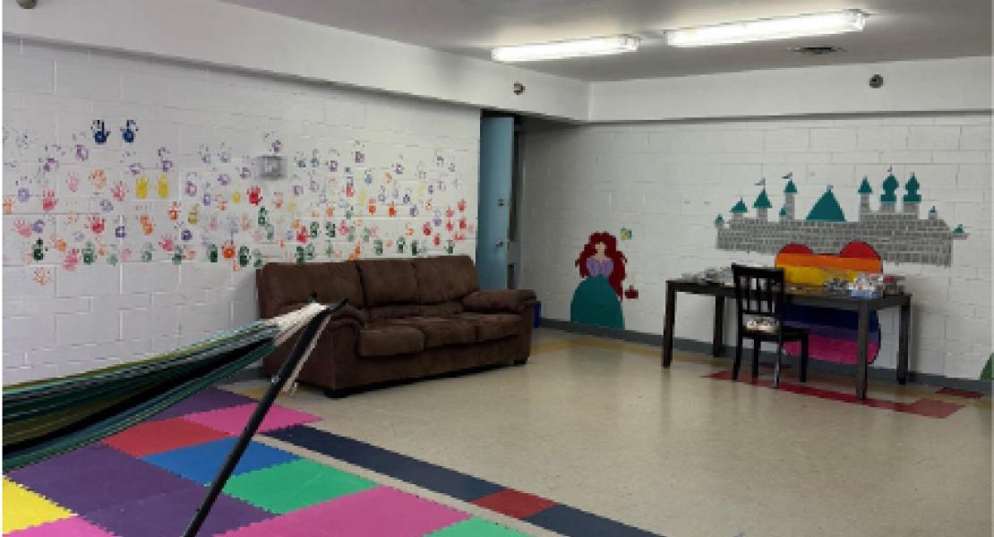 White walled room with bright paintings on the walls, light flooring, colorful foam mats, a hammock, and a brown couch 