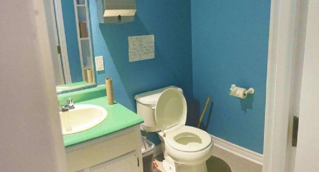Small bathroom with blue walls, green sink counter, mirror, open toilet, wall sign, and trash bin beside sink.