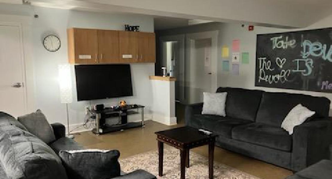 Living room area with light blue walls, patterned area rug, small table, three black couches and a flatscreen tv.  