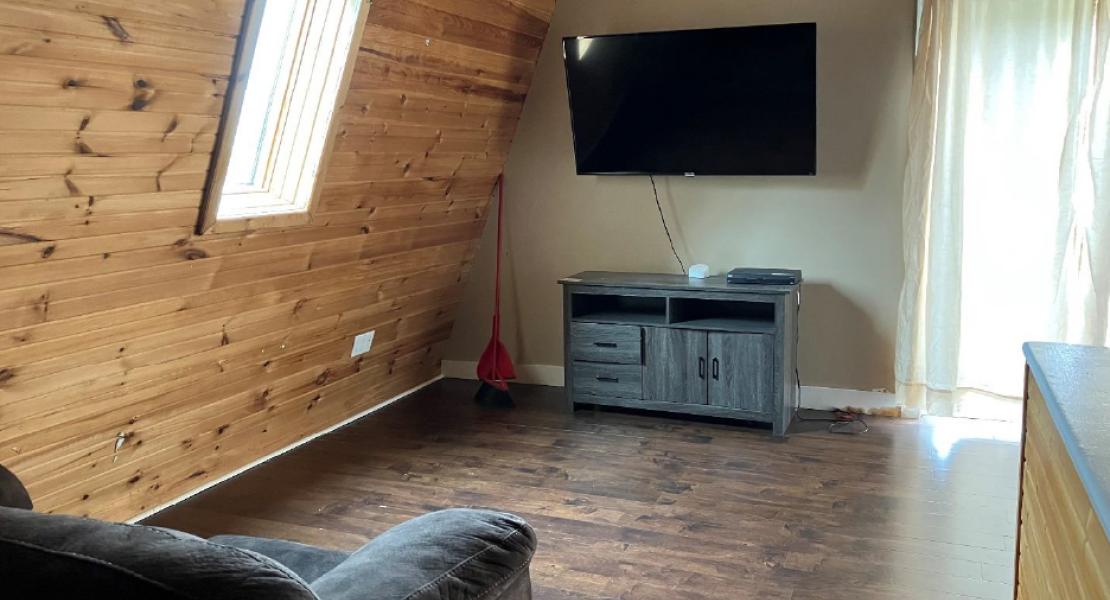 Loft area with dark floors, wooden left wall, gray couch, mounted television, console table, a bright window and patio door leading to an upstairs balcony 