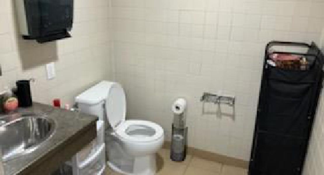 Small bathroom with brown tiled floors and walls, a toilet, shelf area, set of plastic drawers, sink and paper towel dispenser. 