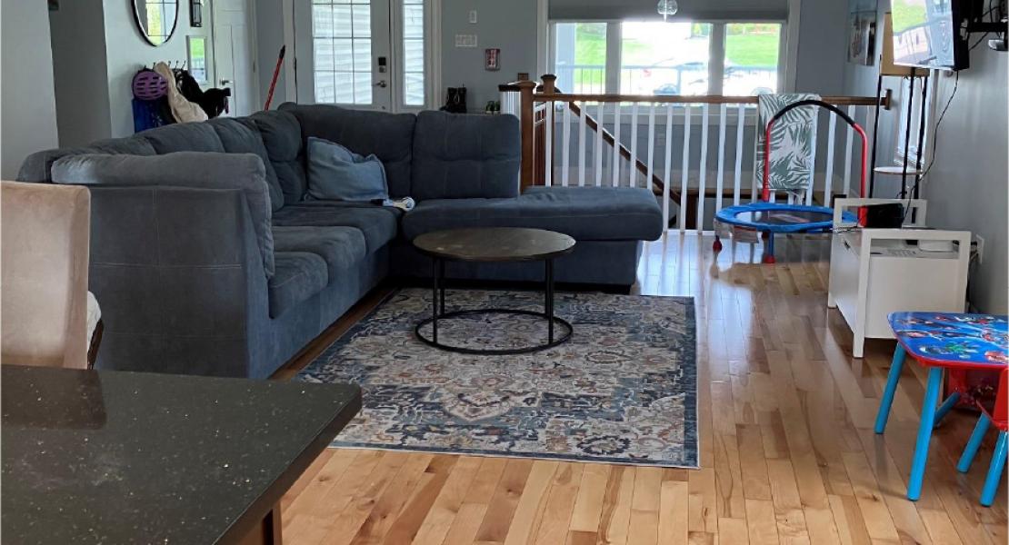 Open living space with hardwood floors, sectional sofa, rug, mounted TV, kids’ table, and indoor mini trampoline near a window 