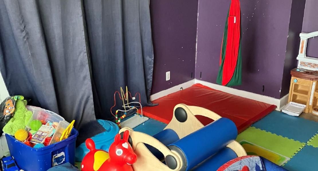 Sensory room with foam mats, toys, a red and green sensory swing and dark curtains against a starry purple wall. 