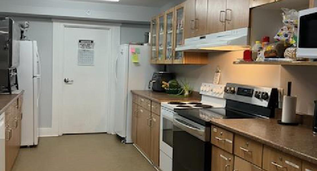 Kitchen area with white door, light brown floors, blue walls, wooden cupboards and a variety of appliances including two stoves and a milk machine. 