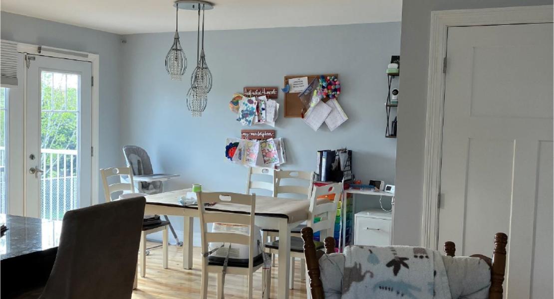  Dining area with light wood flooring, chairs around a table, hanging lights, and children’s artwork displayed on the wall. 