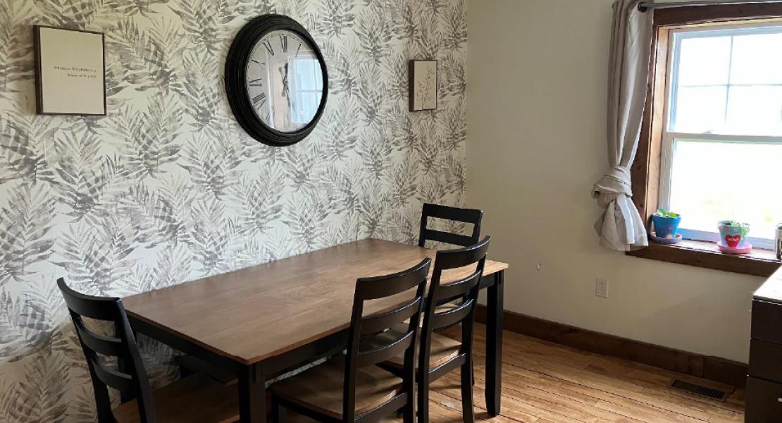 Dining area with wooden floors, a table and four chairs, a bright window, a large clock on a floral wall papered wall 