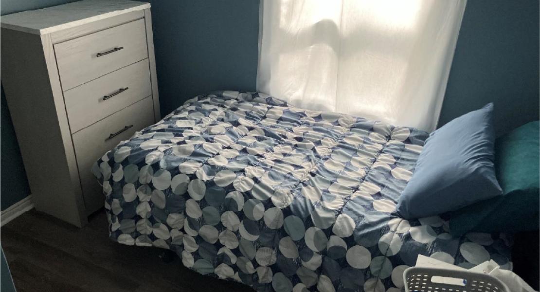 A bedroom with a single bed, blue bedding, white dresser, and window 