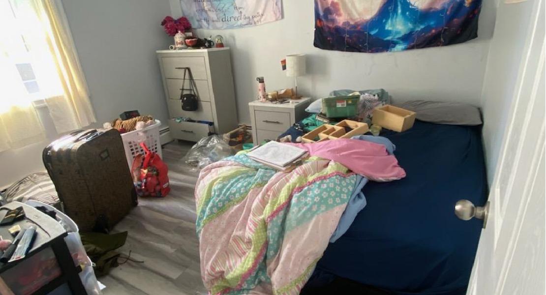 Bedroom with wood floors, dresser, nightstand, suitcase, clothes and bed with dark sheet 