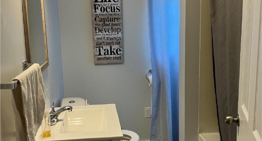 Small bathroom with blue walls, dark flooring, tub with shower curtain, mirror, sink, toilet and window covered with blue curtain 