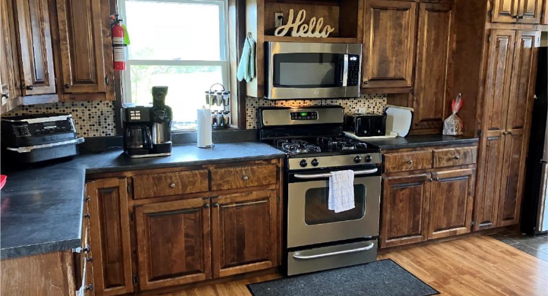 Kitchen area with dark wood cupboards, stainless steel microwave and stove, with one bright window and wooden floors. 