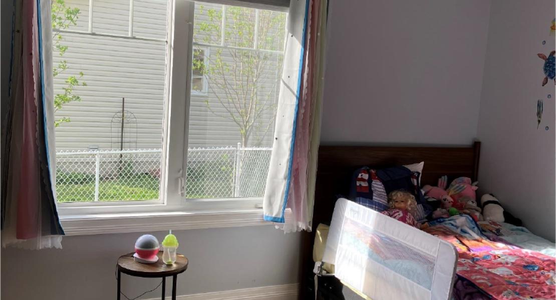 Bedroom with hardwood floors, a single bed with colourful bedding, large side-facing window and a small round table. 